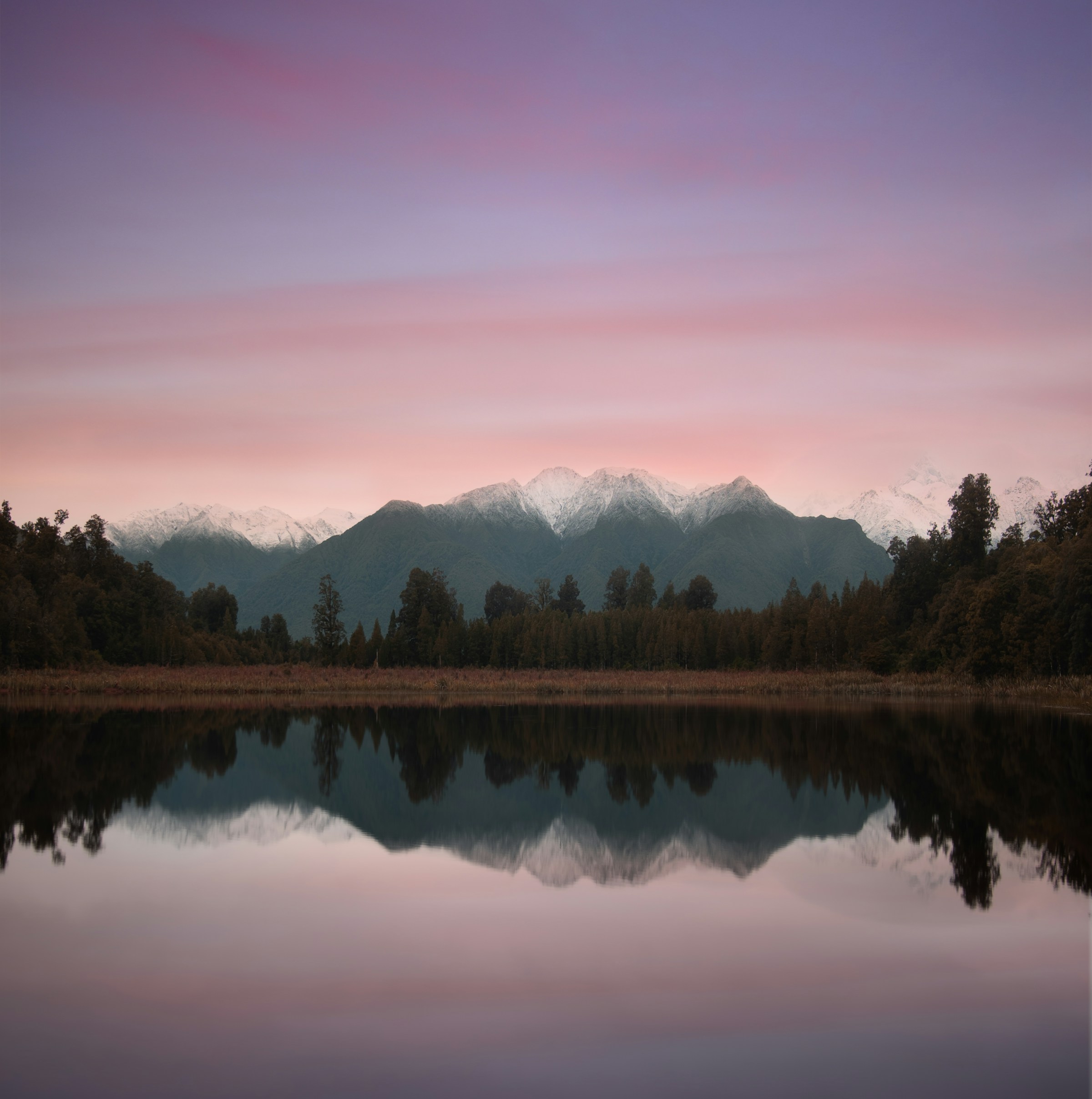 Snow-capped mountains and trees reflected in still water at dusk